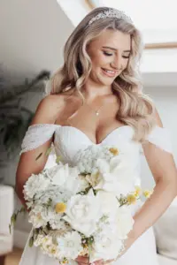 A smiling bride with long blonde hair in loose waves, wearing an off-the-shoulder white wedding dress and jeweled headband, holds a bouquet of white and yellow flowers at her Bunny Hills wedding.