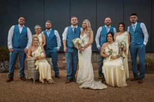 A wedding party poses outdoors at Bunny Hills. The bride stands in the center in a white gown holding flowers, surrounded by bridesmaids in pale yellow dresses and groomsmen in blue vests and ties. Some sit on chairs, while others stand.