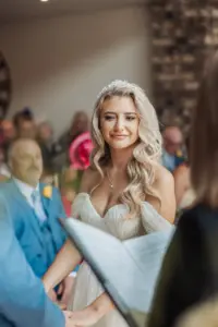 A bride with long blonde hair in an off-the-shoulder white wedding dress smiles warmly while holding hands during a charming Bunny Hills wedding ceremony, with guests seated in the background.