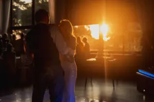 A couple dances closely in a softly lit room at their bunny hills wedding, with warm sunlight streaming through large windows and silhouetted guests watching.