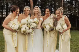 A bride in a white dress happily shows her ring while standing with four bridesmaids in matching light yellow dresses, all holding white flower bouquets outdoors at a beautiful Bunny Hills wedding surrounded by trees.