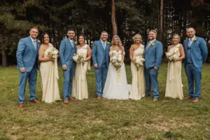 A wedding party poses outdoors at Bunny Hills. The bride and groom stand in the center, surrounded by bridesmaids in light yellow dresses and groomsmen in blue suits, all holding bouquets or wearing boutonnieres, with trees in the background.