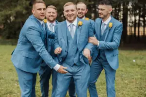 Five men in matching blue suits and yellow boutonnieres stand together outdoors at a Bunny Hills wedding, smiling and laughing. Four playfully crowd around the man in the center, who appears to be the focus of their attention.