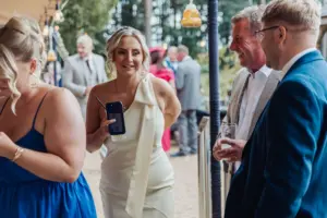 A group of people dressed formally are socializing at a bunny hills wedding. A woman in a white dress holds a phone and smiles, while others around her chat and hold drinks. The background is decorated with string lights and greenery.