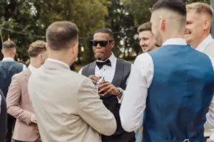A group of men dressed in suits and tuxedos socializing outdoors at a Bunny Hills wedding; one man in sunglasses and a bow tie holds a drink while others face him, with trees and string lights in the background.