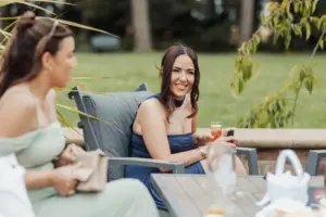 Two women sit outdoors on patio chairs at a Bunny Hills wedding, talking and smiling. One, in a blue dress, holds a drink and looks at the camera. The background features green grass, trees, and blurred foliage at this social gathering.