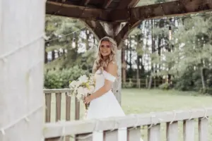 A bride in an off-the-shoulder white gown stands under a wooden gazebo at her Bunny Hills wedding, holding a bouquet of white and yellow flowers, smiling at the camera with trees and greenery visible in the background.