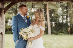 A bride in a white off-the-shoulder dress smiles at her groom in a blue suit with a yellow boutonniere. They stand together outdoors under wooden beams with hanging lights at their beautiful Bunny Hills wedding.