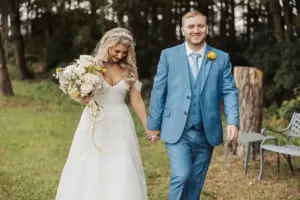A bride in a white dress holding a bouquet walks hand in hand with her groom in a blue suit at their Bunny Hills wedding. Theyre smiling outdoors, surrounded by trees and grass—a perfect scene for saying I do.