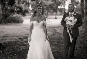 A bride in an off-shoulder wedding dress smiles as she walks outdoors beside a groom holding a bouquet of flowers at a Bunny Hills wedding. The scene, set in a tree-filled area, is beautifully captured in black and white.