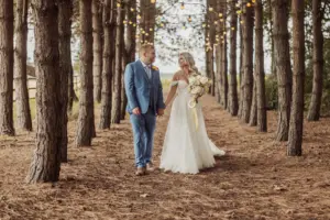 A bride and groom walk hand in hand through a pine tree grove decorated with string lights at their Bunny Hills wedding. The bride wears a white dress and holds a bouquet; the groom wears a blue suit. The setting is rustic and romantic.