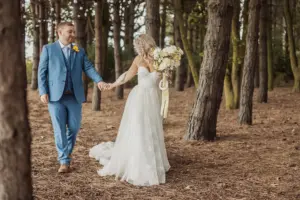 A bride in a white gown and a groom in a blue suit hold hands and walk together through the tall trees of Bunny Hills, creating a magical forest wedding scene. The bride carries a large bouquet of flowers.