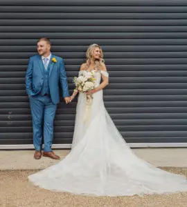 A bride in a white off-shoulder gown smiles at her groom in a blue suit and yellow boutonniere. Hand in hand, they share a joyful moment before a dark metal shutter at their Bunny Hills wedding.