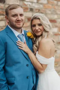A bride in an off-shoulder white dress stands close to a groom in a blue suit with a yellow boutonniere. Both are smiling softly, posing against a rustic brick wall at their Bunny Hills wedding.