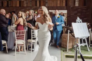 A smiling bride in a white off-shoulder dress walks into her Bunny Hills wedding reception as guests seated at tables applaud and smile. A seating chart on an easel stands nearby.