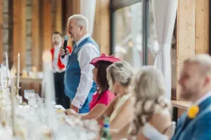 A man in a blue vest and white shirt stands and speaks into a microphone at a long banquet table, while people seated nearby listen attentively during a warmly lit bunny hills wedding in a rustic venue.