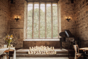 A stone-walled room at Barden Tower with a large arched window, filled with natural light. Numerous white candles line the floor. A black grand piano and a bouquet of flowers on a table create an elegant wedding ambiance.