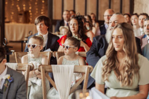 Wedding guests seated indoors at Barden Tower, including two young girls in white dresses and heart-shaped sunglasses in the foreground, surrounded by formally dressed adults, all facing forward and listening attentively.