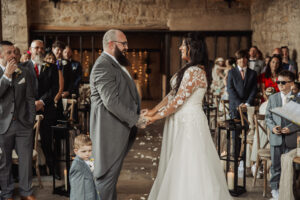 A bride and groom stand holding hands, smiling at each other during a wedding ceremony indoors at the Priests House. Guests sit and stand in the background, while a young boy in a suit stands before the groom looking at the camera.