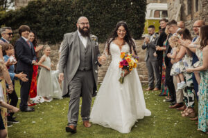 A newlywed couple walks hand-in-hand outdoors at the Priests House, Barden Tower, smiling as guests celebrate on both sides. The bride holds a vibrant bouquet; the groom is in a gray suit. Flower petals are scattered on the grass.