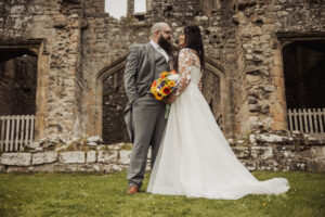 A bride in a white gown and a groom in a gray suit stand close, smiling and holding a colorful bouquet, in front of the historic Barden Tower with its arched windows—the perfect setting for a romantic wedding near the Priests House.
