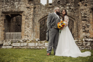 A bride in a white dress holding a bouquet of sunflowers stands beside a groom in a grey suit at Barden Tower’s ancient stone ruins, both looking into the distance on their wedding day.