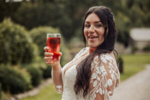 A smiling woman in a white lace wedding dress holds up a glass of red drink outdoors at the Priests House, with lush greenery and blurred trees around this charming Barden Tower wedding venue.