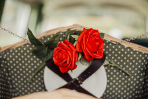Two vibrant red roses with green leaves rest on a wicker basket with a polka dot lining, beside leather straps and white plates, creating a romantic wedding picnic setting near Barden Tower.