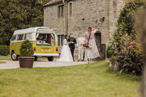 A bride and groom stand with two others outside the Priests House at Barden Tower, posing for a wedding photo beside a vintage green van while a woman in a pink dress takes their picture on a sunny day.
