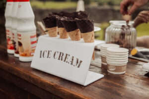 Chocolate-dipped waffle cones are displayed in a white metal holder labeled ICE CREAM on a wooden counter at Barden Tower, perfect for a wedding reception at the Priests House, with cups, bottles, and a glass jar in the background.