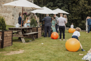 A group of people in formal clothes gather outdoors under white umbrellas near wooden tables at Barden Tower. Children play with large orange exercise balls on the grass, with trees and the old priests house stone wall in the background.