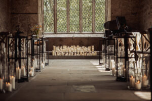 A stone aisle lined with lanterns and candles leads to a large window; many candles are arranged at the end of the aisle, creating a warm, cozy wedding atmosphere in the historic, rustic setting of Barden Tower.