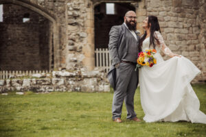 A smiling bride and groom stand on grass in front of the stone ruins of Barden Tower. The groom wears a gray suit, while the bride, holding a colorful bouquet, lifts her dress—both joyfully posing at their unforgettable wedding.