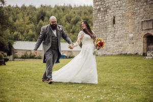 A bride in a white dress holding a bouquet and a groom in a grey suit hold hands and smile as they walk on grass outside, celebrating their wedding near the Priests House at Barden Tower, with trees and a stone building in the background.
