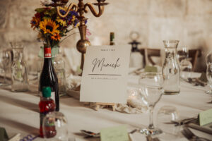 A wedding table setting with a sign reading Table Five Munich in elegant script, surrounded by flowers, wine bottles, glassware, candles, and green place cards on a white tablecloth at the charming Barden Tower Priests House.