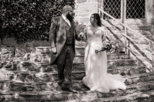 A bride and groom holding hands and walking down the stone steps outside Barden Tower, with the bride in a long gown holding a bouquet and the groom in a suit and glasses. Sunlight casts shadows on the steps and wall at this romantic wedding venue.