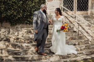 A bride in a white dress holding a colorful bouquet walks down stone steps, hand in hand with her bearded groom in a grey suit, outside the historic Barden Tower Priests House at their wedding, framed by ivy and leaded windows.