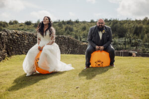 A bride in a white dress and a groom in a suit joyfully bounce on large orange space hoppers outdoors at the Priests House, Barden Tower, with a grassy lawn, stone wall, and green trees highlighting their playful wedding moment.