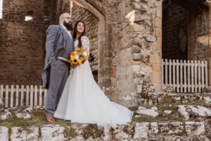A bride in a white dress and a groom in a gray suit stand together on stone steps, holding a vibrant bouquet, framed by the historic Priests House at Barden Tower and its charming arched doorway.