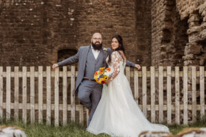 A bride in a white dress and groom in a gray suit stand by a wooden fence at Barden Tower, with the historic Priests House behind them. The bride holds a colorful bouquet with sunflowers, completing their charming wedding scene.