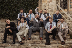 A group of eleven men dressed in suits and ties sit on the stone steps outside the Priests House at Barden Tower, smiling and making playful hand gestures, with a historic stone building and greenery in the background—capturing joyful wedding moments.