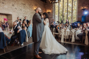 A bride and groom dance together on a black dance floor at their wedding reception in the rustic stone-walled Priests House at Barden Tower, surrounded by seated guests and elegant decorations.