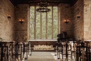 A rustic stone hall at Barden Tower is set up for a wedding, with rows of chairs facing a large window. Candles glow at the front, and a black grand piano sits to one side. Warm wall sconces light the room in the historic Priests House.