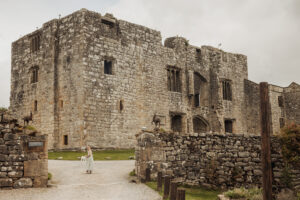 A young girl in a white dress stands near the entrance of Barden Tower, a weathered stone castle with tall walls and small windows under a cloudy sky. Two goat statues are perched on the stone wall by the path, evoking a wedding near the ancient priests house.