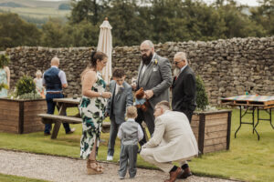 A group of adults and children in formal attire gather on the grass near picnic tables and a stone wall at Barden Tower. One man kneels, talking to a small boy in a suit, as others interact in this relaxed wedding setting.