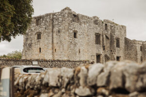 A weathered stone castle, known as Barden Tower, with tall, narrow windows stands behind a low stone wall and parked cars, surrounded by greenery under a cloudy sky—a remarkable setting often chosen for a wedding or linked to the former priests house.