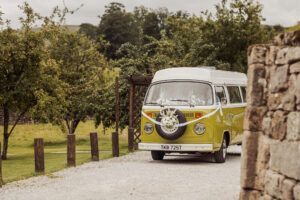 A vintage yellow and white van decorated with flowers and ribbons drives on a gravel path by Barden Tower’s stone wall, near the Priest’s House, surrounded by green trees on a cloudy wedding day.