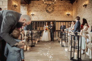 A bride in a white dress walks down the aisle with an older man in a suit. Guests watch as flower petals and candles line the aisle of the rustic stone Priests House at Barden Tower, creating a magical wedding atmosphere.