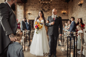 A bride in a white gown holding a colorful bouquet walks down the aisle of the rustic, stone-walled Barden Tower wedding venue with a man in a suit. Guests watch as candles line the aisle. A child and a bearded man stand at the front.