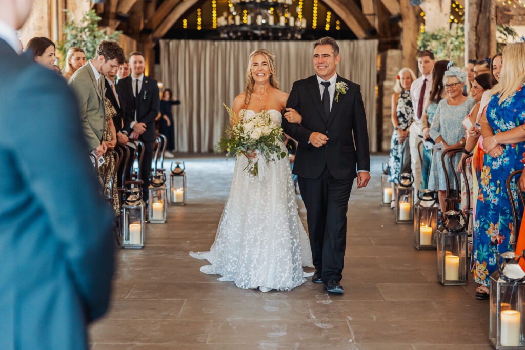 A smiling bride in a white dress walks arm-in-arm with a man in a black suit down an aisle decorated with lanterns, surrounded by seated guests in a warmly lit venue.